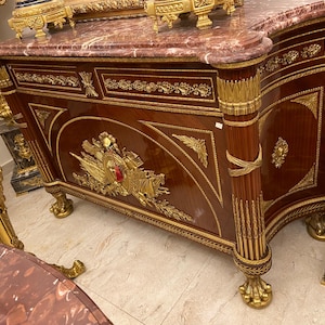 May include: A mahogany-toned antique cabinet with ornate gold accents. The cabinet features a marble top with red veining, decorative gold trim, and detailed carvings. The legs are gold-colored and claw-shaped, adding to the antique aesthetic.