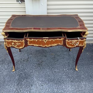 May include: A vintage desk with a dark brown leather top and three drawers. The desk has a mahogany finish with gold accents and ornate carvings on the legs and drawer fronts. The desk is a classic piece of furniture.