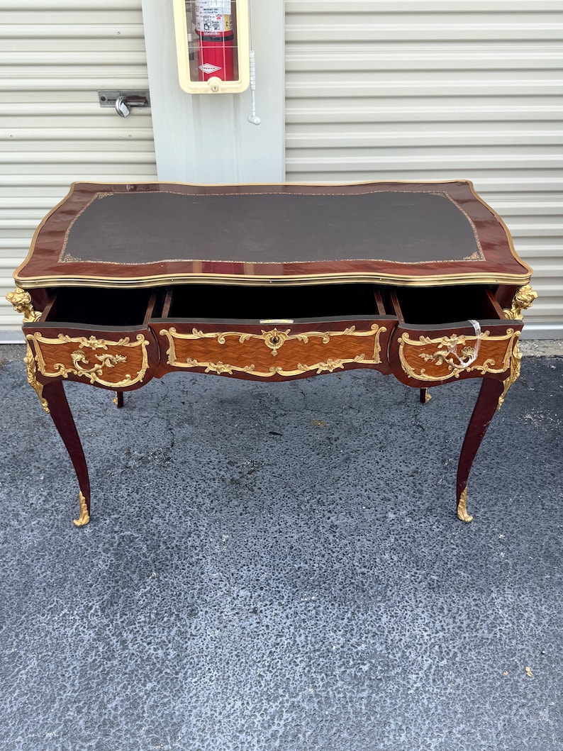 May include: A vintage desk with a dark brown leather top and three drawers. The desk has a mahogany finish with gold accents and ornate carvings on the legs and drawer fronts. The desk is a classic piece of furniture.
