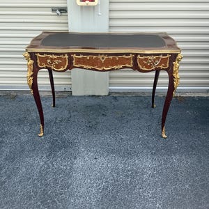 May include: An antique desk with a dark brown leather top and gold trim. The mahogany desk features ornate gold accents on the legs and drawers. The curved legs and overall design give it a classic, elegant appearance. The desk is approximately 120 cm long.