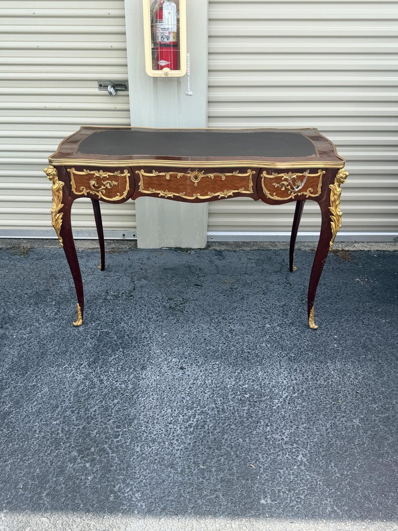 May include: An antique desk with a dark brown leather top and gold trim. The mahogany desk features ornate gold accents on the legs and drawers. The curved legs and overall design give it a classic, elegant appearance. The desk is approximately 120 cm long.