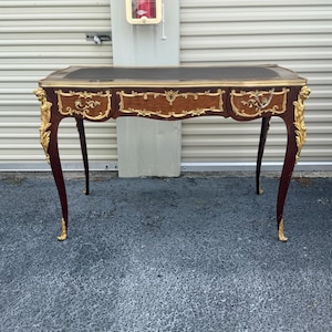 May include: A mahogany desk with a black leather top and three drawers. The desk has ornate gold accents on the legs and drawer fronts. The desk's curved legs and decorative details give it a classic, elegant appearance.