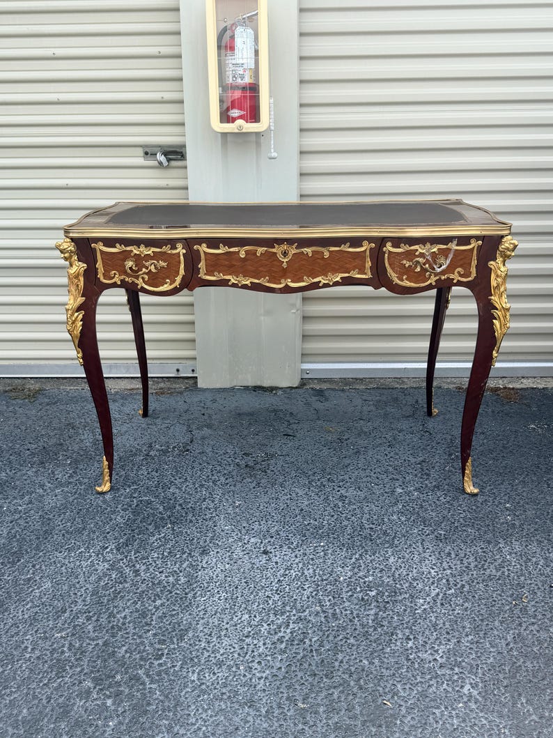 May include: A mahogany desk with a black leather top and three drawers. The desk has ornate gold accents on the legs and drawer fronts. The desk's curved legs and decorative details give it a classic, elegant appearance.