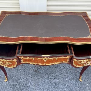 May include: A mahogany desk with a dark brown leather top and three drawers. The desk has ornate gold accents and curved legs. The drawers are open, revealing the interior. The desk is a classic piece of furniture.