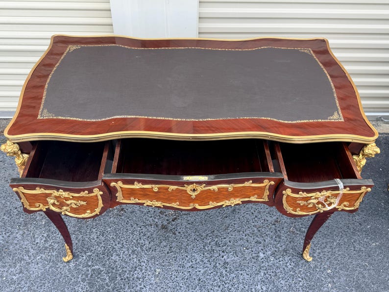 May include: A mahogany desk with a dark brown leather top and three drawers. The desk has ornate gold accents and curved legs. The drawers are open, revealing the interior. The desk is a classic piece of furniture.