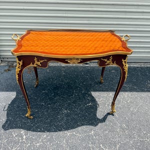 May include: An antique side table with a rectangular top featuring an orange patterned surface and a dark wood frame. The table has ornate gold accents, curved legs, and gold handles. A classic piece of furniture.