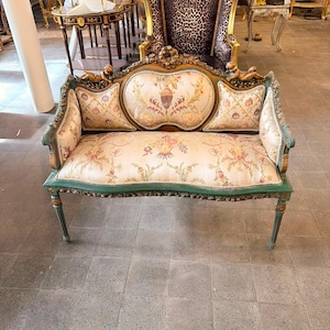 May include: A settee featuring a green and gold painted wooden frame with ornate carvings and two small cherub figures. The upholstery is a cream-colored fabric decorated with a multi-colored floral pattern and a central urn motif on the backrest.