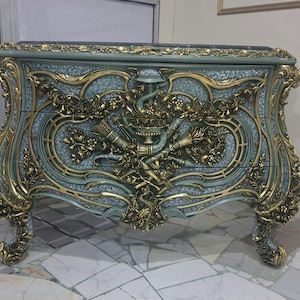 May include: A chest of drawers featuring a light blue speckled finish with ornate gold-colored relief carvings. The front displays three drawers decorated with intricate floral motifs, scrolls, and a central emblem, supported by four curved legs.