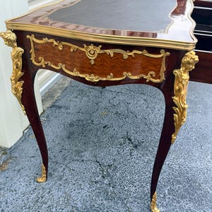 May include: An antique desk with a dark brown leather top and ornate gold accents. The desk features a mahogany finish with intricate carvings and curved legs. The desk is approximately 76 cm tall and 122 cm wide.