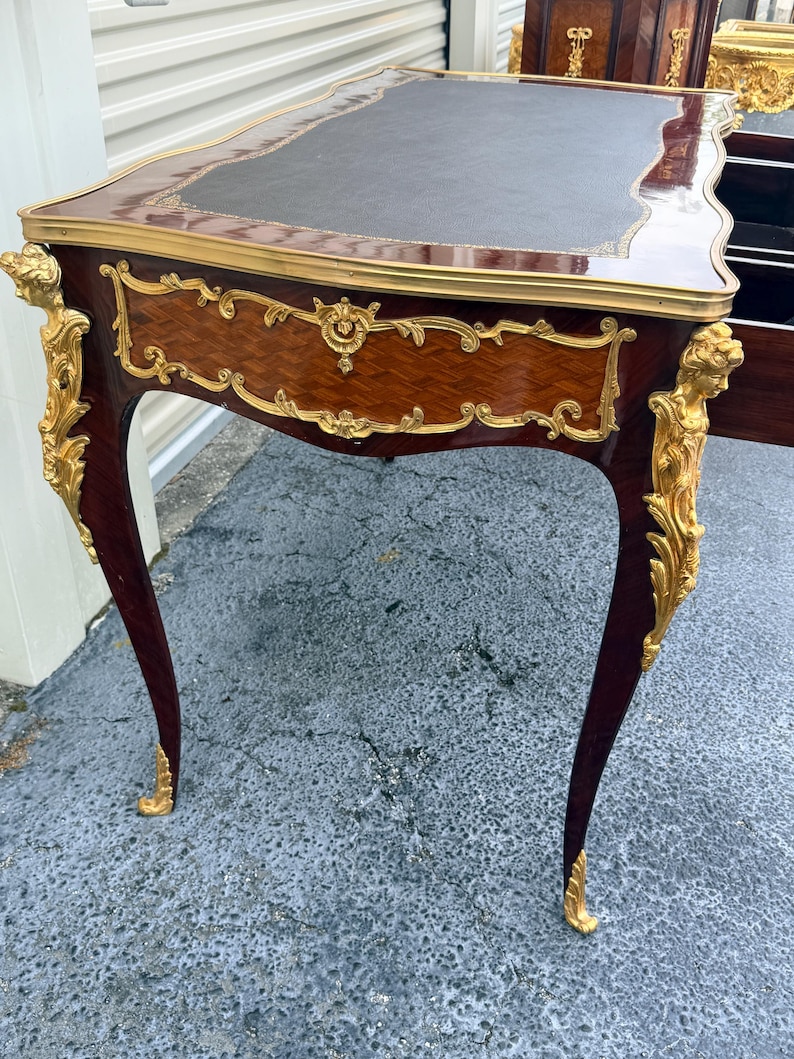 May include: An antique desk with a dark brown leather top and ornate gold accents. The desk features a mahogany finish with intricate carvings and curved legs. The desk is approximately 76 cm tall and 122 cm wide.