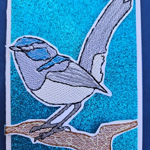 May include: Embroidered bird art on a teal background. The bird is primarily gray with blue accents, perched on a brown branch. The artwork is on a white card with a teal glittery background.
