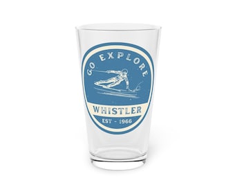May include: A clear glass pint with a blue and white graphic. The graphic features a skier with the words "GO EXPLORE" and "WHISTLER EST - 1966". The glass is empty and sits on a white surface.