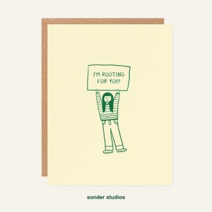 May include: A greeting card with a light yellow background and a brown border. The card features a green illustration of a person holding a sign that reads "I'M ROOTING FOR YOU!". The words "sonder studios" are printed at the bottom.