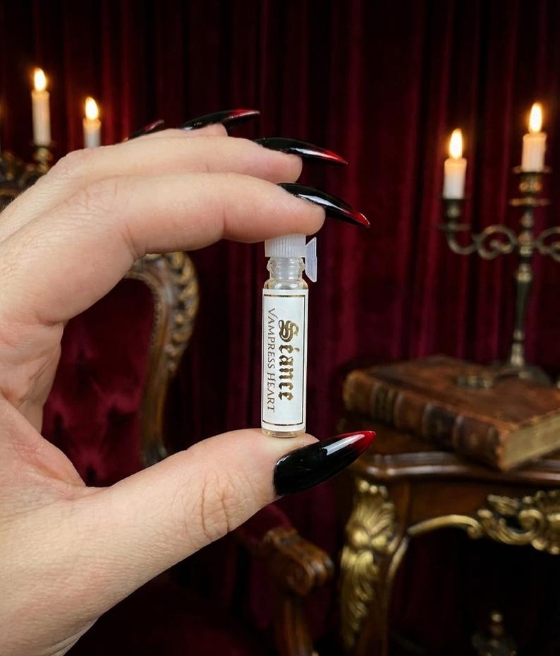 May include: A small, clear glass vial with a white plastic stopper and a rectangular white label. The label features the text 'S&eacute;ance' in a decorative font above the words 'VAMPRESS HEART' in a simple, capitalized sans-serif typeface.