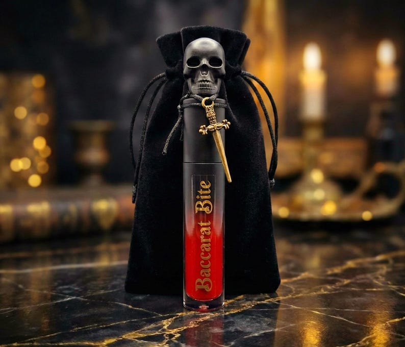 May include: A black lipstick tube with the text "Baccarat Bite" in gold lettering, a gold sword charm, and a black skull charm. The lipstick is in front of a black velvet drawstring bag. The lipstick has a red tint.