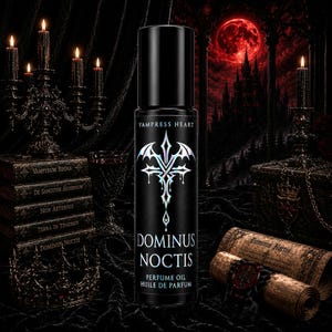 May include: A black cylindrical perfume oil bottle labeled VAMPRESS HEART and DOMINUS NOCTIS in white text, featuring a silver holographic emblem of a stylized cross with bat-like wings above the words PERFUME OIL HUILE DE PARFUM.