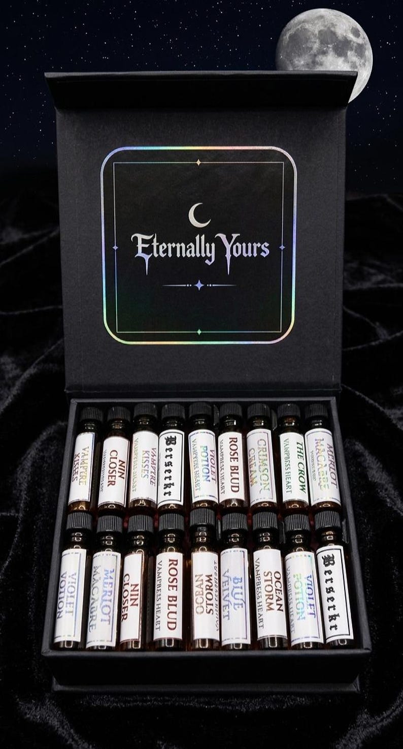 May include: A black rectangular box containing sixteen small glass vials with black caps. Each vial features a white label with text including names like Berserker, Rose Blud, and Crimson Cream. The box lid displays the text Eternally Yours in white.