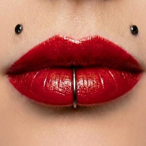 May include: Close-up of glossy red lips with a silver lip ring piercing the lower lip. Two black piercings are visible above the upper lip. The skin is smooth and the lips are full and well-defined.