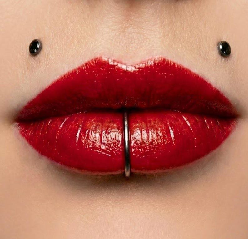 May include: Close-up of glossy red lips with a silver lip ring piercing the lower lip. Two black piercings are visible above the upper lip. The skin is smooth and the lips are full and well-defined.