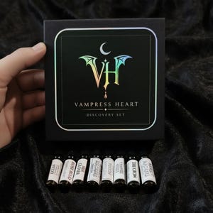 May include: A black square box labeled VAMPRESS HEART DISCOVERY SET with a holographic logo featuring a crescent moon and bat wings. Below the box are eight small glass vials with white labels, each containing a different fragrance name.