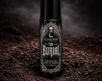 May include: A black cylindrical perfume oil bottle with a label featuring a white gothic arch, a skull, a crescent moon, and the text VAMPRESS HEART THE BURIAL PERFUME OIL HUILE DE PARFUM. The bottle stands upright with a smooth, glossy finish.