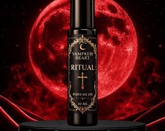 May include: A black cylindrical perfume oil bottle with a label featuring gold ornate borders, a crescent moon, a cross, and the text VAMPRESS HEART RITUAL PERFUME OIL 10 ML. The bottle has a black cap and is centred against a large red moon.