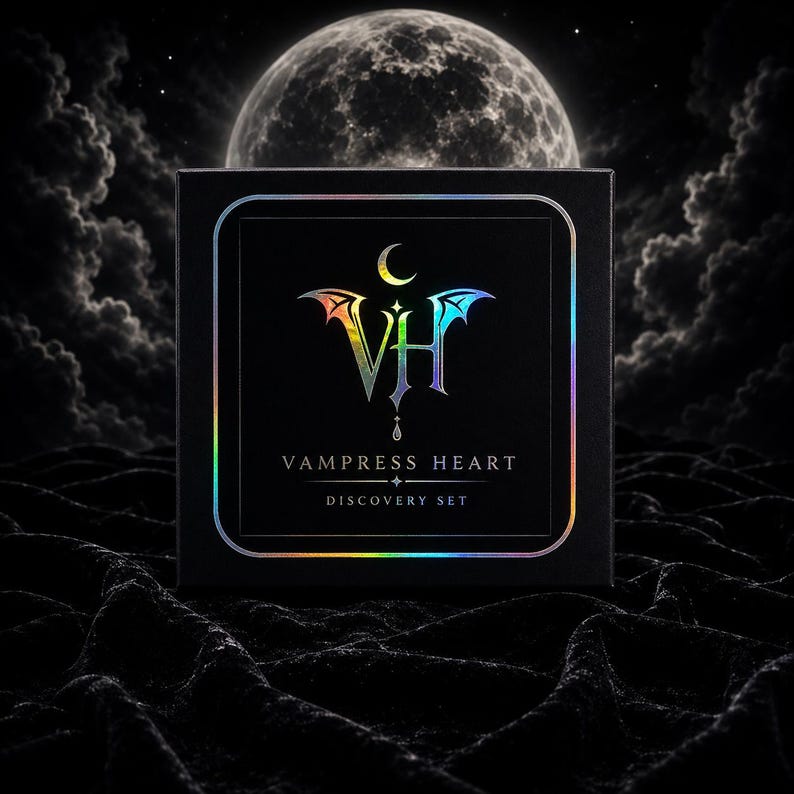 May include: A square black box featuring a holographic logo with the letters VH, bat wings, and a crescent moon. Below the logo, the text reads VAMPRESS HEART and DISCOVERY SET. The box is framed by a thin holographic border on a black surface.