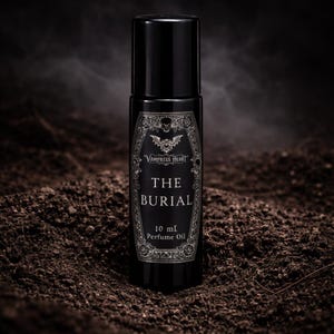 May include: A black perfume oil bottle with a label that reads "Vampress Heart THE BURIAL 10 ml Perfume Oil." The label features a decorative frame with a bat and skull design. The bottle is set against a dark, earthy background.