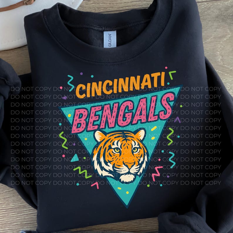 May include: Black sweatshirt featuring the text "Cincinnati Bengals" in a retro design. The design includes a tiger head graphic inside a teal triangle, with colorful confetti accents. A Gildan tag is visible.