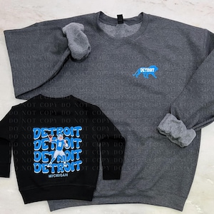 May include: Two sweatshirts, one black and one heather grey. The black sweatshirt has a graphic of a football player with the text "DETROIT" repeated multiple times and "MICHIGAN" at the bottom. The grey sweatshirt has a blue lion graphic with the text "DETROIT" above it.