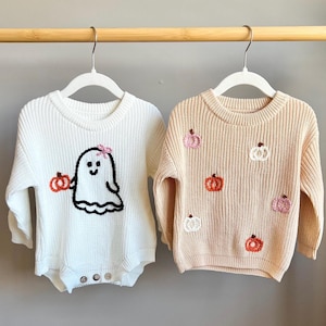 May include: Two children's sweaters hanging on a rack. One is white with a ghost and pumpkin design. The other is tan with embroidered pumpkins. Both sweaters have long sleeves and are made of a knitted material.