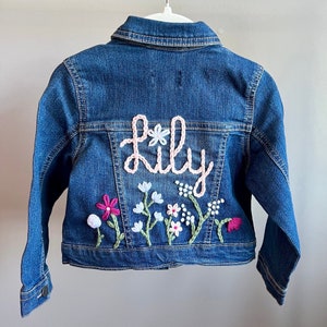 May include: A blue denim jacket with the name "Lily" embroidered in pink cursive script. The back of the jacket features embroidered flowers in pink, white, and blue, with green stems and leaves. The jacket is hanging on a white hanger.