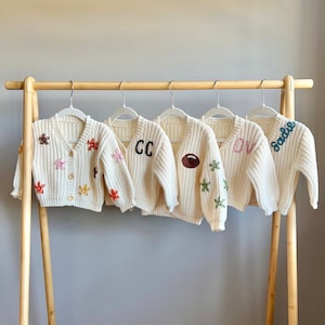 Peut inclure: Cinq cardigans en tricot blanc avec des détails brodés. Les cardigans présentent différents motifs, notamment des fleurs, des lettres et un ballon de football.