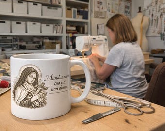 May include: White ceramic mug featuring a sepia-toned illustration of a religious figure holding a cross and flowers, with Latin text. The mug is on a wooden table with sewing tools and a woman using a sewing machine in the background.