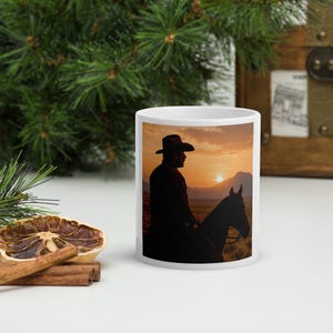 Könnte beinhalten: Weiße Keramik-Tasse mit einer Silhouette eines Cowboys auf einem Pferd vor einer Sonnenuntergangskulisse. Der Cowboy trägt einen Hut und reitet ein Pferd. Der Hintergrund umfasst eine Bergkette und eine untergehende Sonne. Die Tasse hat eine Standardgröße.
