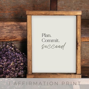 May include: A framed print with the words "Plan. Commit. succeed" in black and gray font. The frame is made of dark brown wood. The print is on a cream-colored background. The words "1 Affirmation Print" are at the bottom.