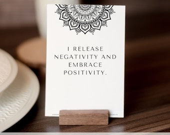 May include: A white card with a black mandala design at the top and the text "I release negativity and embrace positivity." The card is standing on a wooden stand.