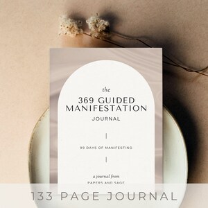 May include: A white journal with the text "the 369 GUIDED MANIFESTATION JOURNAL" and "99 DAYS OF MANIFESTING". The journal is on a beige surface with a white plate and dried flowers.