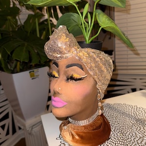 Peut inclure: Un pot de fleurs décoratif en forme de tête avec un turban à paillettes dorées, un maquillage dramatique et un col en velours. Le pot contient une petite plante verte avec de grandes feuilles. La palette de couleurs est marron, or et rose.