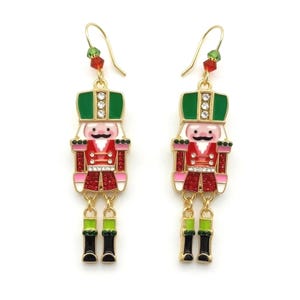 May include: A pair of gold-tone dangle earrings featuring nutcracker figures with green hats, red jackets, and black boots. The nutcrackers are accented with clear rhinestones and small green and red beads on the ear wires, creating a festive look.