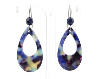 Pode incluir: Um par de brincos em forma de gota com um padrão marmorizado em tons de azul, roxo e amarelo. Cada brinco tem uma conta azul escura e um gancho prateado. Os brincos são exibidos em um suporte transparente.