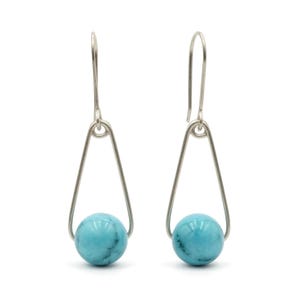 May include: A pair of drop earrings featuring a polished, round turquoise bead suspended within a silver-toned triangular wire frame. The earrings include a curved silver-toned ear wire hook for attachment, showcasing a simple, symmetrical design.