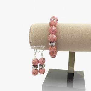 May include: A jewelry set featuring a bracelet and earrings. The bracelet and earrings are made with pink and white marbled beads, separated by silver-colored spacers. The jewelry is displayed on a beige stand against a white background.