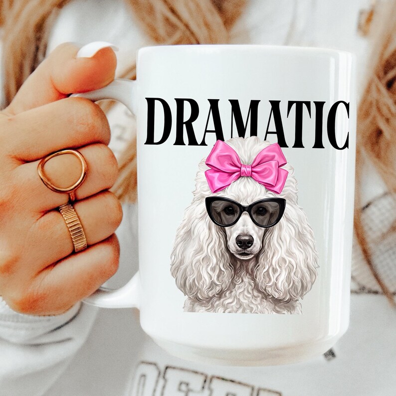 May include: White ceramic mug featuring the word "DRAMATIC" in black capital letters. Below the text is a graphic of a white poodle wearing black sunglasses and a pink bow. The mug is held by a person wearing gold rings.