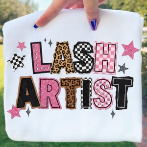 Peut inclure: Sweat-shirt blanc avec l'inscription "LASH ARTIST" dans un design coloré et audacieux. Les lettres présentent des imprimés léopard, des motifs à carreaux, du rose et du noir, avec des étoiles. Le sweat-shirt est tenu par une main.
