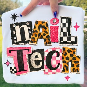 May include: White t-shirt with the words "NAIL TECH" in a colorful, textured design. The letters feature leopard print, glitter, and checkered patterns, with rhinestone and star accents. The design has a distressed, torn paper effect.