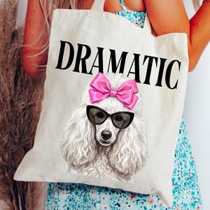 May include: A cream-colored tote bag with the word "DRAMATIC" in large black letters. The bag features a graphic of a white poodle wearing black sunglasses and a pink bow. The poodle is centered on the bag.