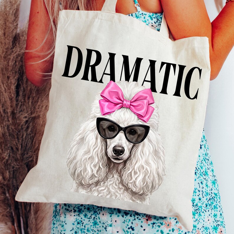 May include: A cream-colored tote bag with the word "DRAMATIC" in large black letters. The bag features a graphic of a white poodle wearing black sunglasses and a pink bow. The poodle is centered on the bag.