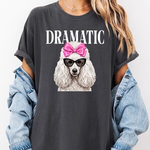 May include: Dark gray t-shirt featuring the word "DRAMATIC" in white block letters above a white poodle illustration. The poodle wears black sunglasses and a pink bow. The shirt has short sleeves.