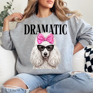 May include: A gray sweatshirt with the word "DRAMATIC" in black block letters. The sweatshirt features a graphic of a white poodle wearing black sunglasses and a pink bow. The poodle's fur is detailed and fluffy.